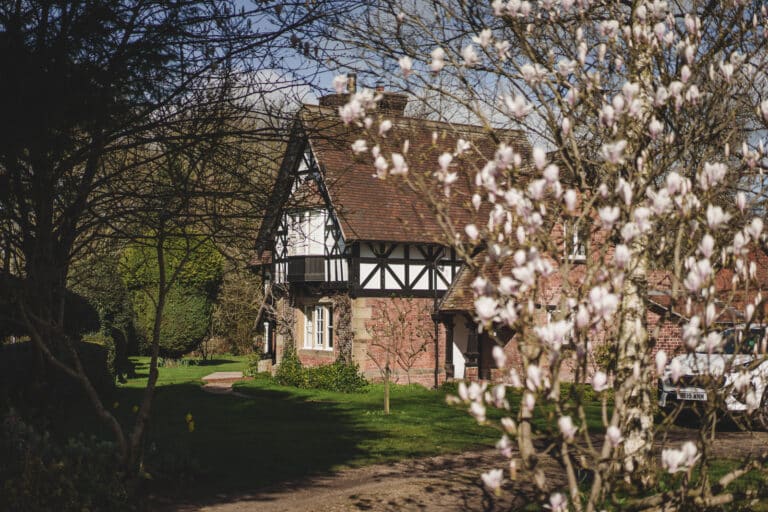 A half-timbered house with a red roof stands behind blooming magnolia trees, surrounded by greenery on a sunny day. A gravel path leads to the house, perfect for a charming wedding..