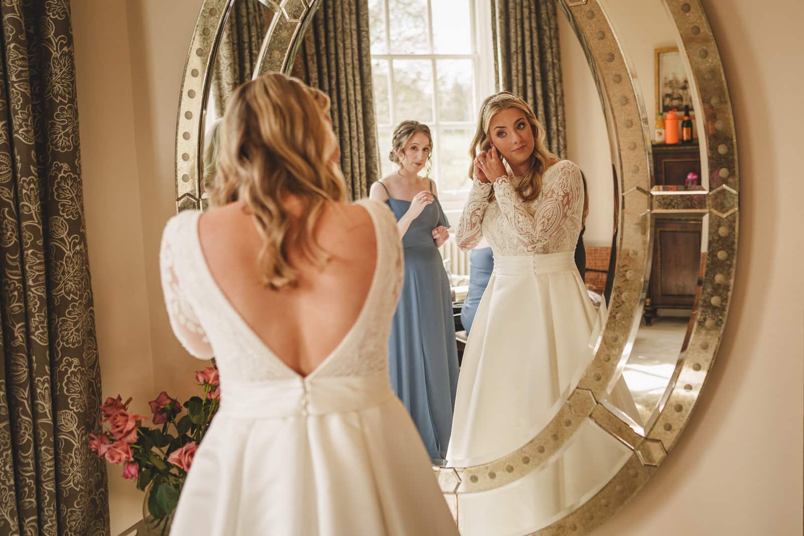 Two women prepare in front of a large mirror. One wears a long-sleeved wedding dress, adjusting her earring, while the other in a blue dress stands behind her. They are in a well-lit room with large windows at Iscoyd Park, Shropshire.