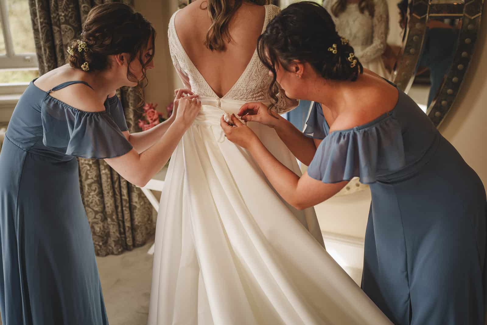 In a well-lit room at the enchanting Iscoyd Park in Shropshire, two bridesmaids in blue dresses help the bride tie the back of her white wedding gown.
