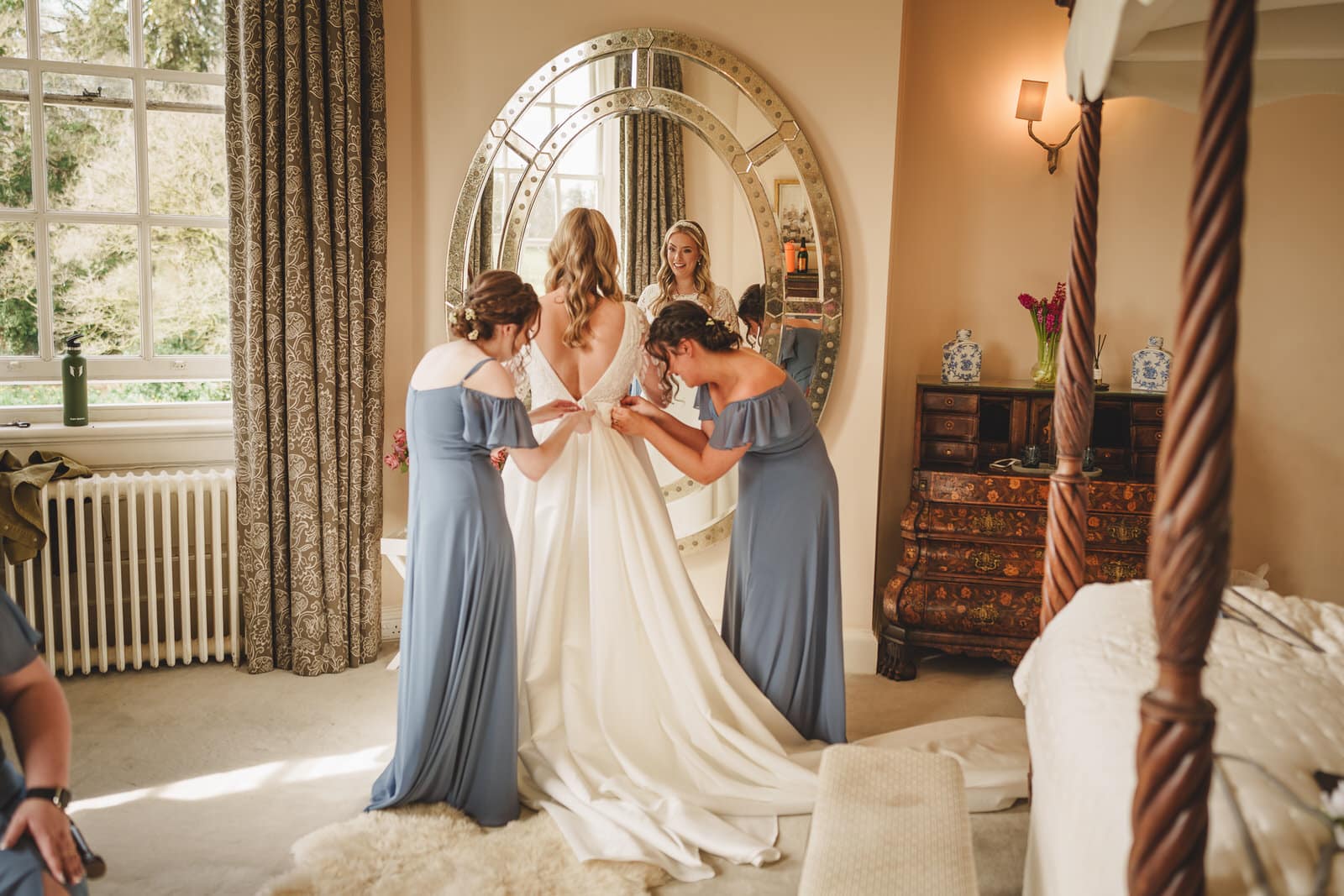 A bride in a white gown stands in front of a large round mirror, assisted by three bridesmaids wearing blue dresses, in a well-furnished room with a window and decorative furniture at Iscoyd Park in Shropshire.