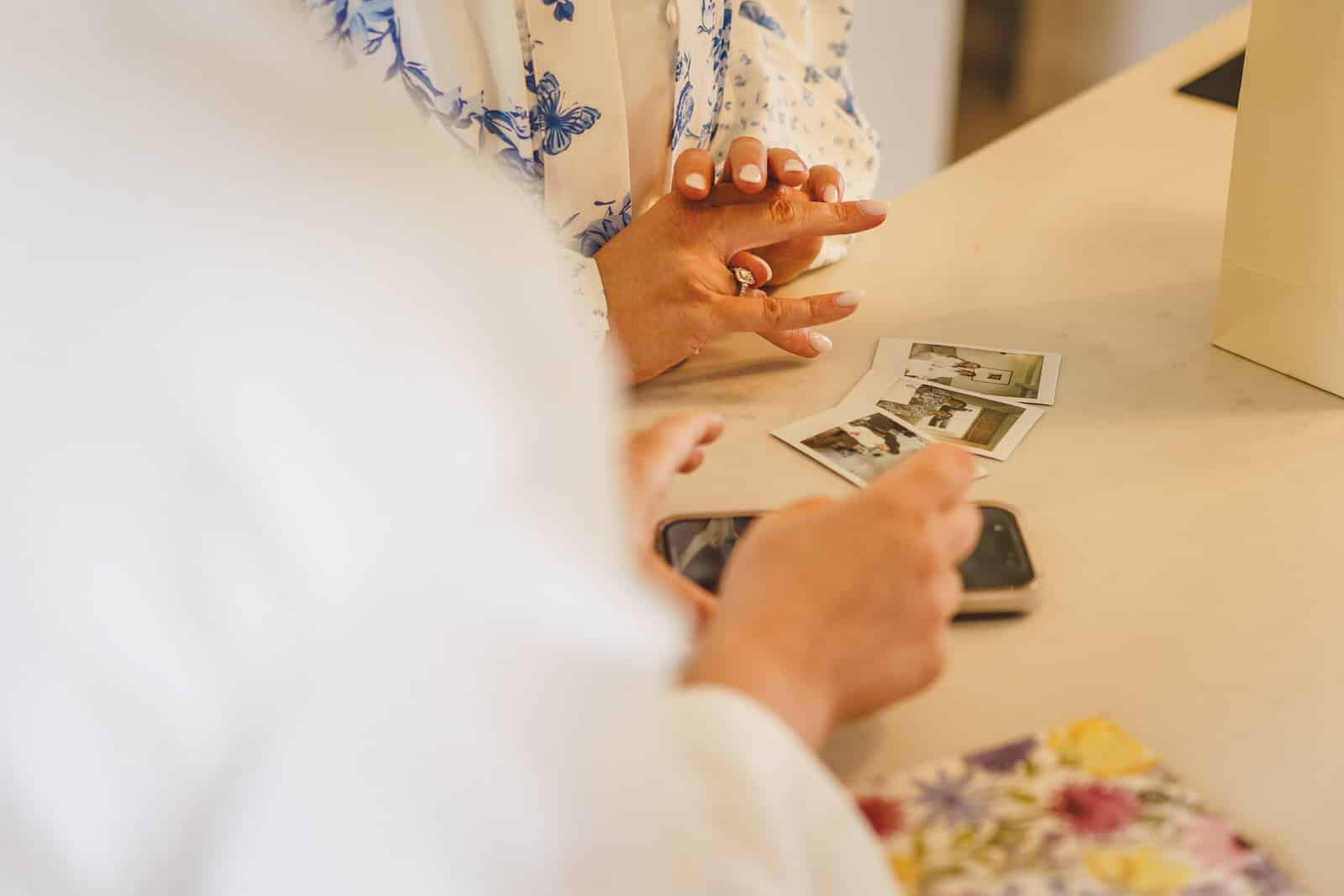 Hamish and Georgia are looking at printed photos on a table. Hamish is holding a smartphone while Georgia rests her hands on the table, both partially visible.