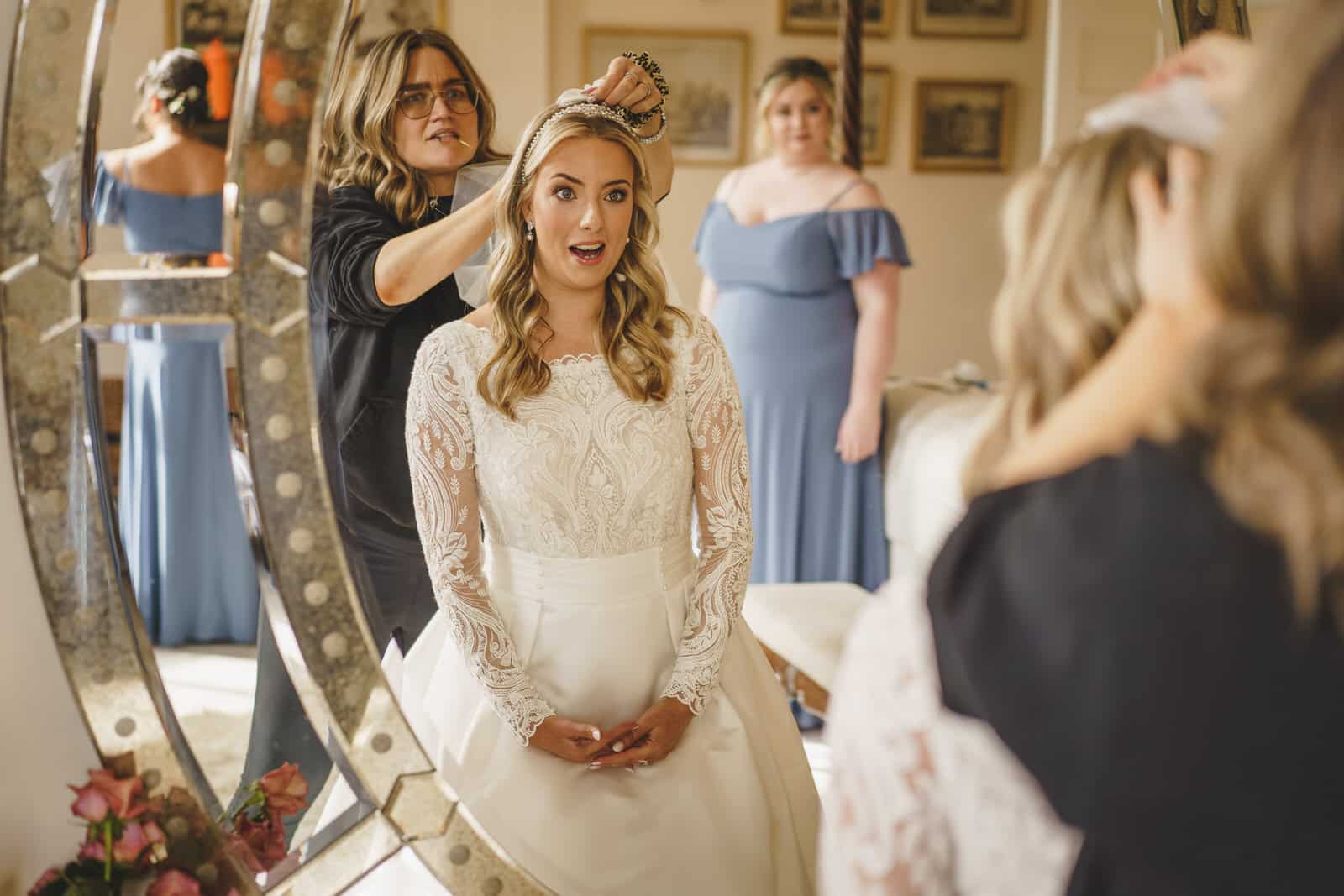 A bride, wearing a lace dress, sits in front of a mirror as a woman places a tiara on her head. Another woman in a blue dress stands in the background at Iscoyd Park, nestled in the charming Shropshire countryside.
