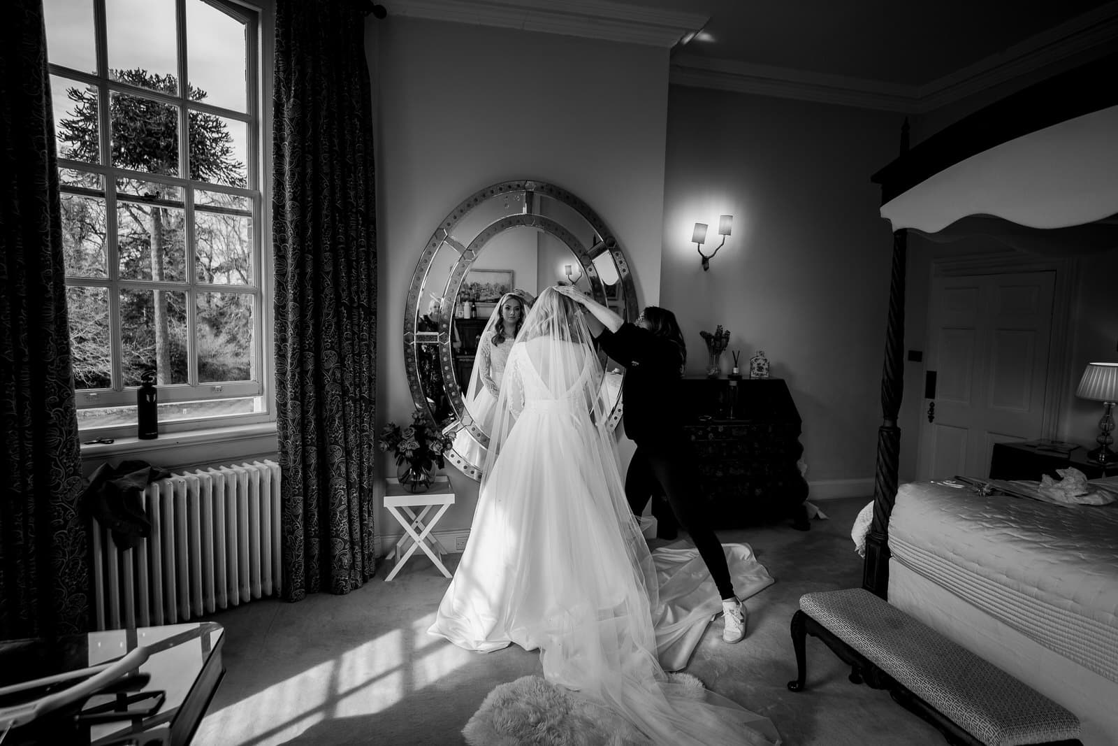 A bride stands in a room facing a large mirror at Iscoyd Park, wearing a wedding dress and holding a bouquet, while an assistant adjusts her veil.