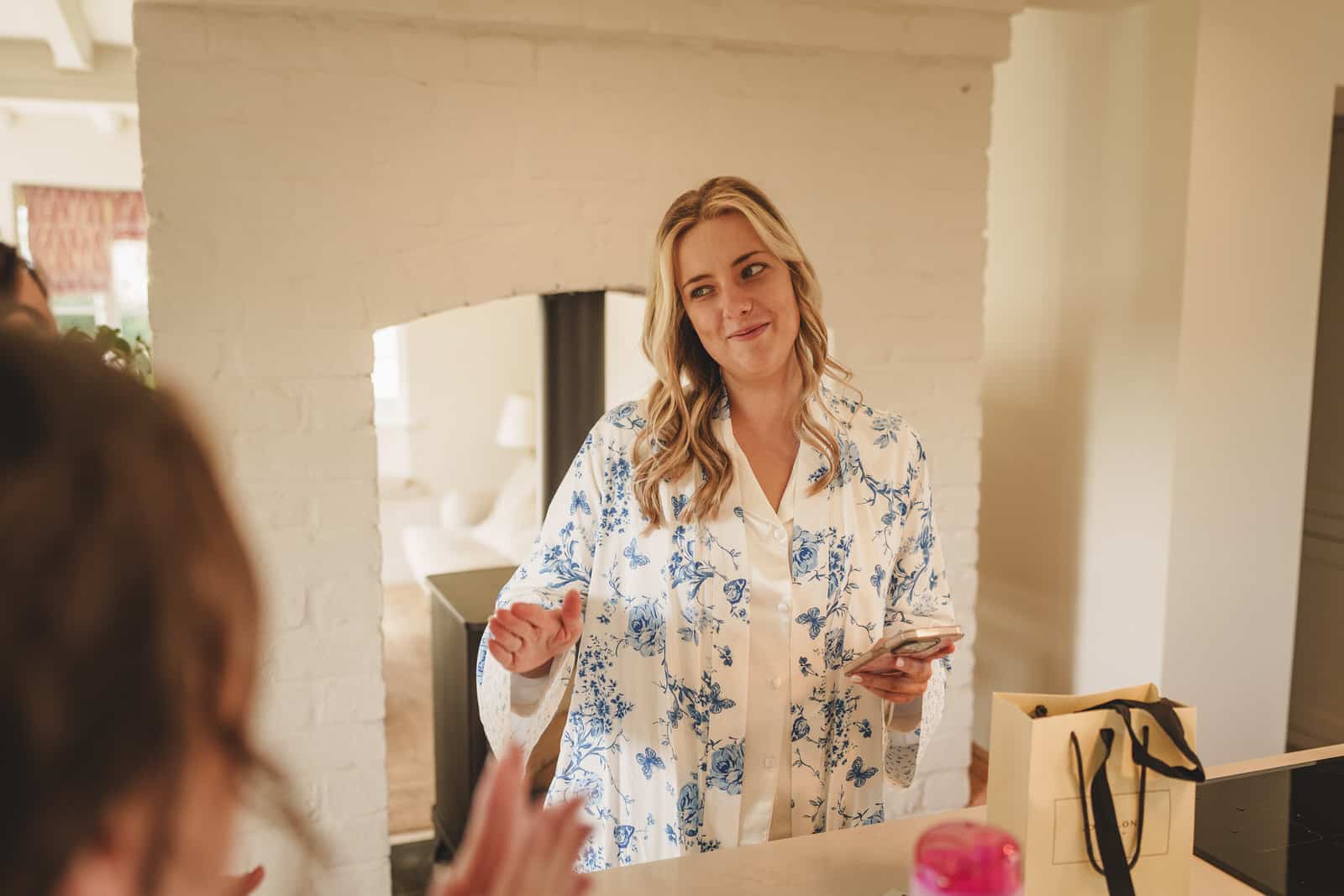 a person wearing a floral patterned robe holds a phone and gestures while standing in a room with a white brick wall and archway, perhaps discussing details for the charming wedding.