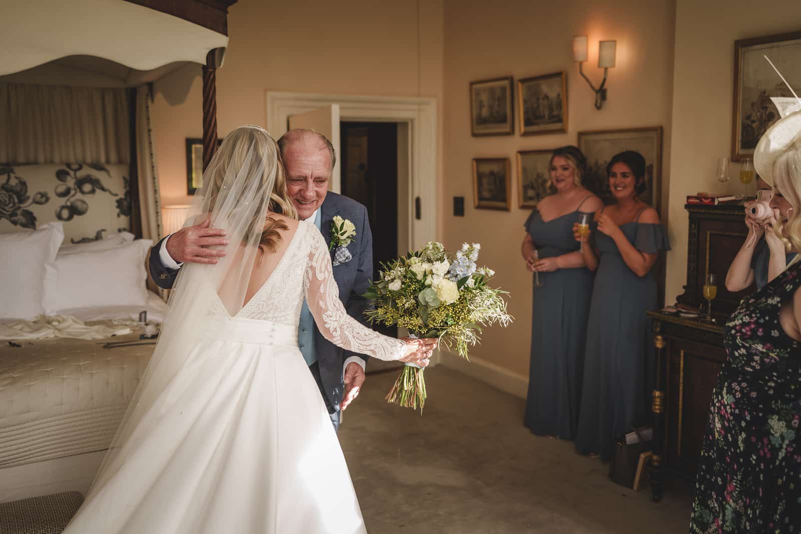 A bride in a white dress and veil hugs her Father in a suit and boutonnière in a bedroom at Iscoyd Park, Shropshire, while three women in matching off-shoulder dresses look on, holding drinks and smiling.