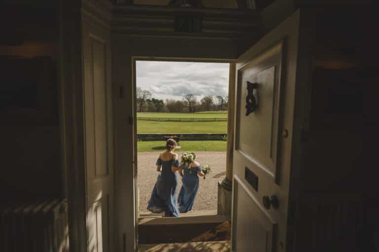 Two people in blue dresses holding flower bouquets walk outside, viewed through an open door with the picturesque Shropshire countryside fields in the background.