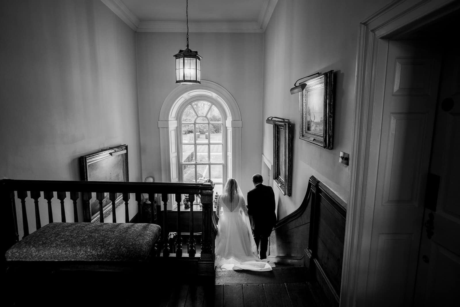 The bride and her Father descend a wide staircase together in a spacious room with a large window in the background, captured in black and white. This charming wedding scene is filled with timeless elegance.