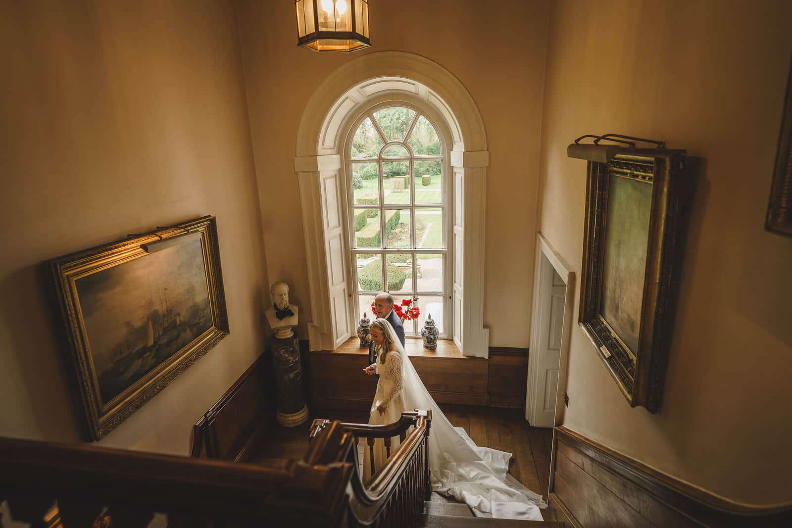 The charming bride and her Father, walk up a wooden staircase in a historical building adorned with framed paintings and a bust. A large arched window with garden views provides a picturesque backdrop for their wedding day.