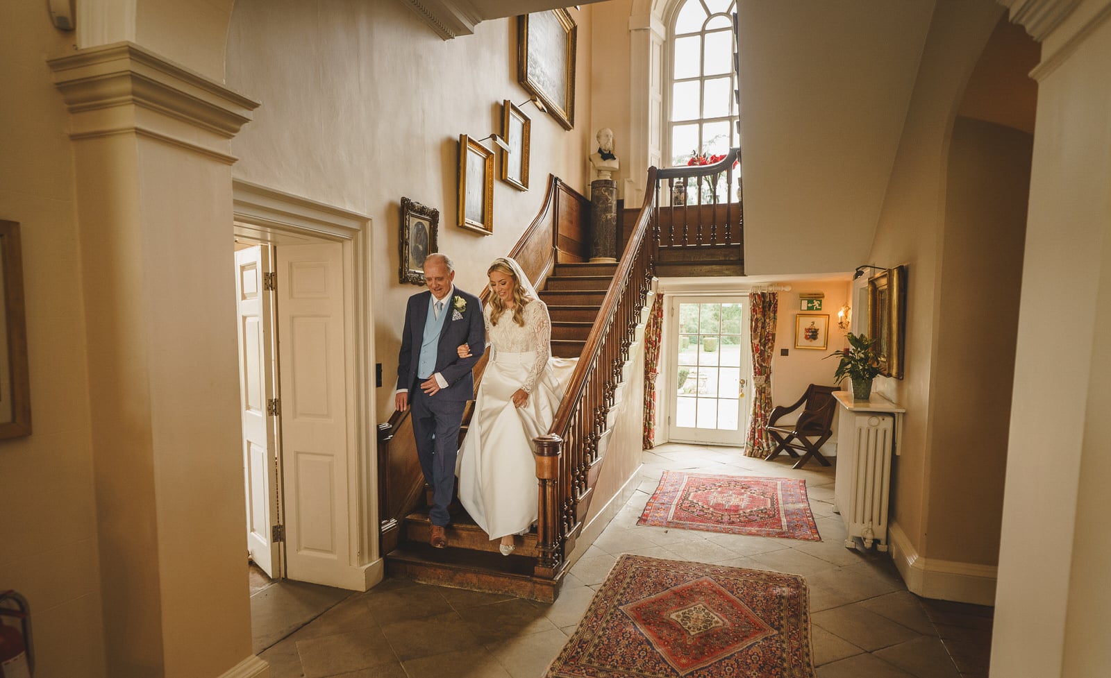 A bride in a white dress descends a staircase with her Father in a blue suit during the wedding ceremony in a well-lit, elegantly decorated hallway with paintings and rugs, capturing the essence of Georgia's rich heritage.