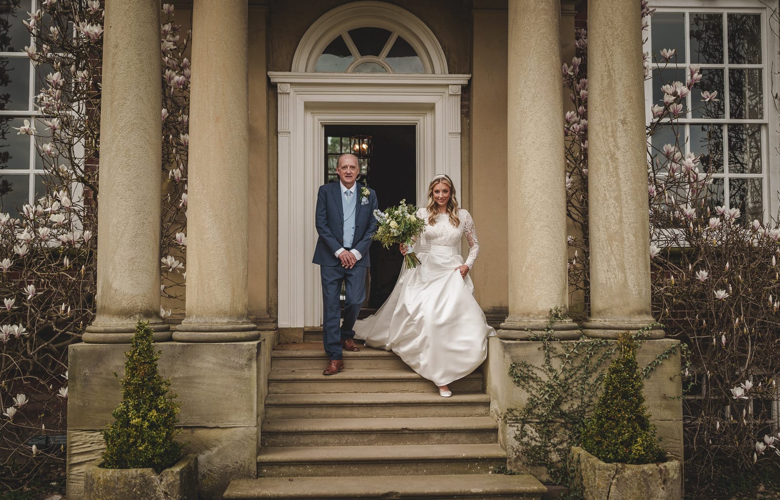 A bride and her father, stand on the steps outside a grand building with large columns and blooming flowers. She is holding a bouquet and wearing a white dress, painting the perfect scene of a charming wedding in Shropshire.