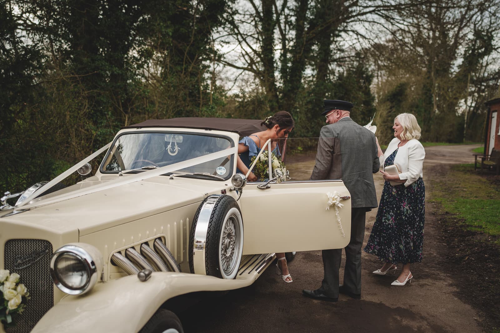 a bride leans into a vintage car adorned with flowers and ribbons at hamish and georgia's charming wedding. an older man and woman stand nearby in a wooded outdoor setting.