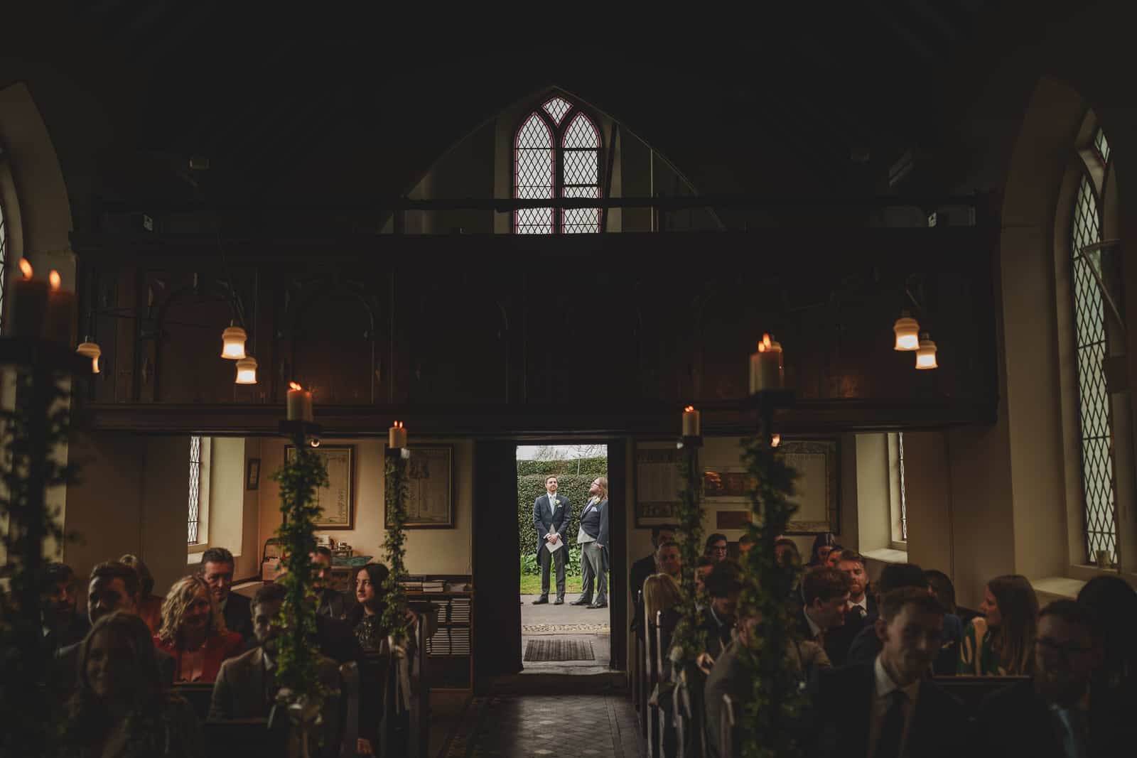 a dimly lit church interior with guests seated in pews. two men, hamish and a friend, stand outside the open door at the back, visible in the georgia sunlight. the aisle is decorated with greenery and candles, setting the scene for a charming wedding.