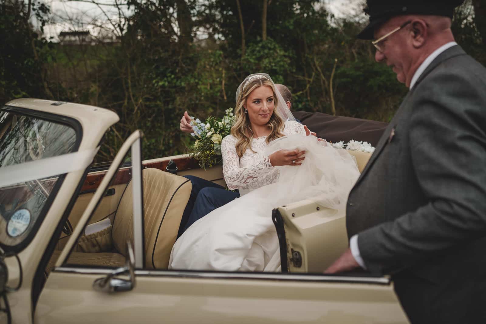 a bride in a white dress and veil sits in the back of a vintage convertible car while being assisted by an older man in a suit and hat. the car's door is open, with greenery in the background, capturing hamish and georgia's charming wedding day.