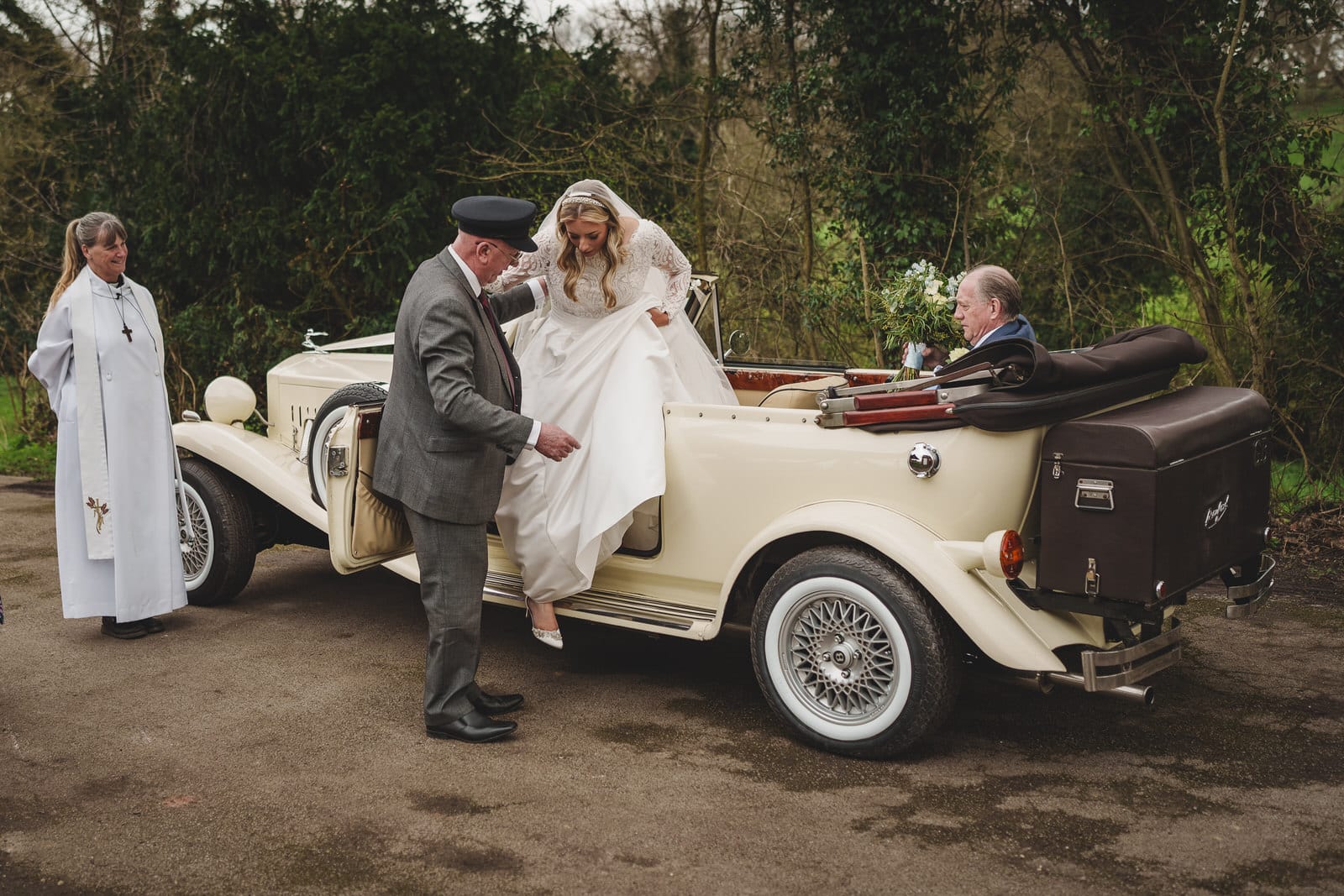 a bride steps out of an ivory vintage car with the assistance of an older man in a suit, while the charming wedding officiant and older man seated in the car, observe the scene.