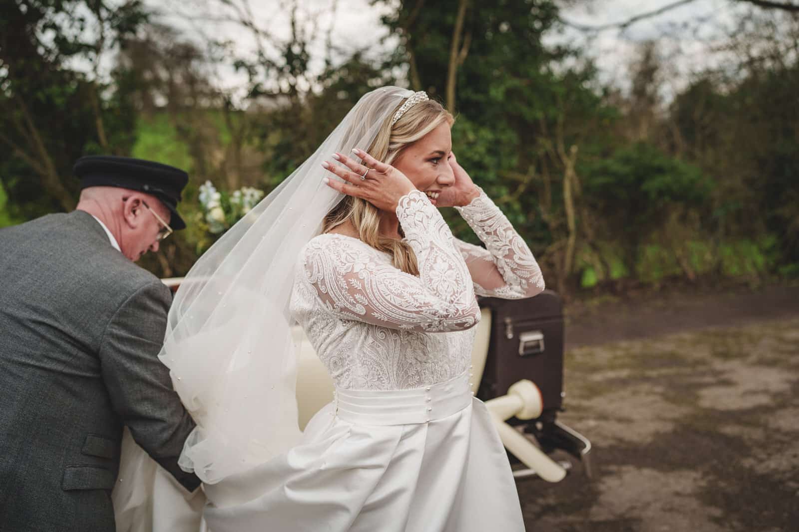 georgia, adjusts her veil while standing outside next to a car. a man, possibly a chauffeur, assists her with the gown. trees and bushes are visible in the background of this charming wedding scene.
