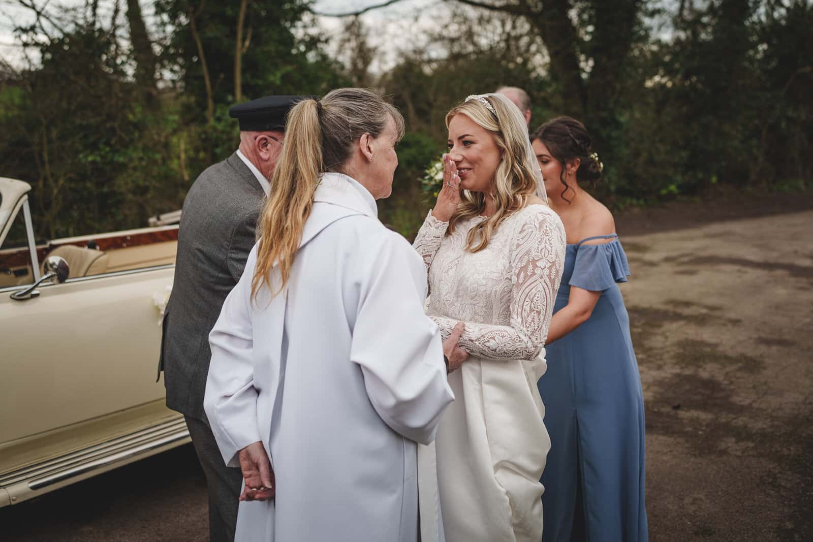 at a charming wedding in shropshire a bride in a white dress stands beside a woman in a white coat, with others around. there’s an old fashioned car in the backdrop and a bridesmaid in a blue dress. trees are in the background.