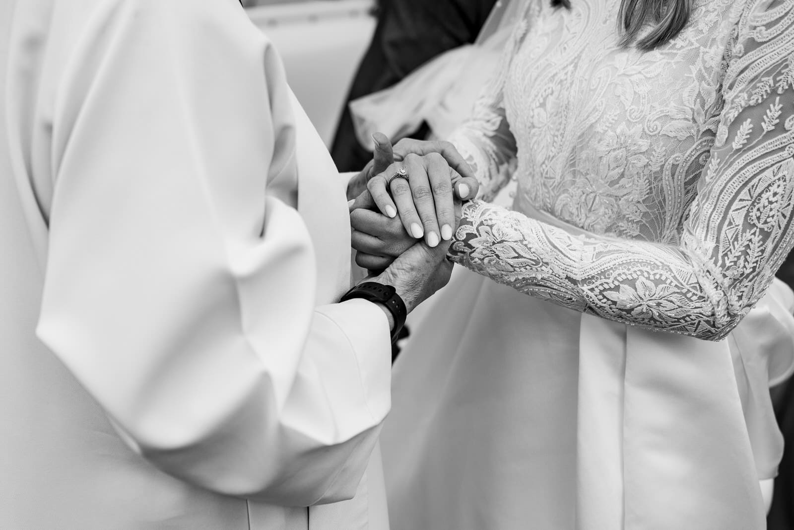 a close up black and white photo captures the essence of a charming wedding as two people hold hands; one is in a lace wedding dress, the other in a light colored robe or suit.
