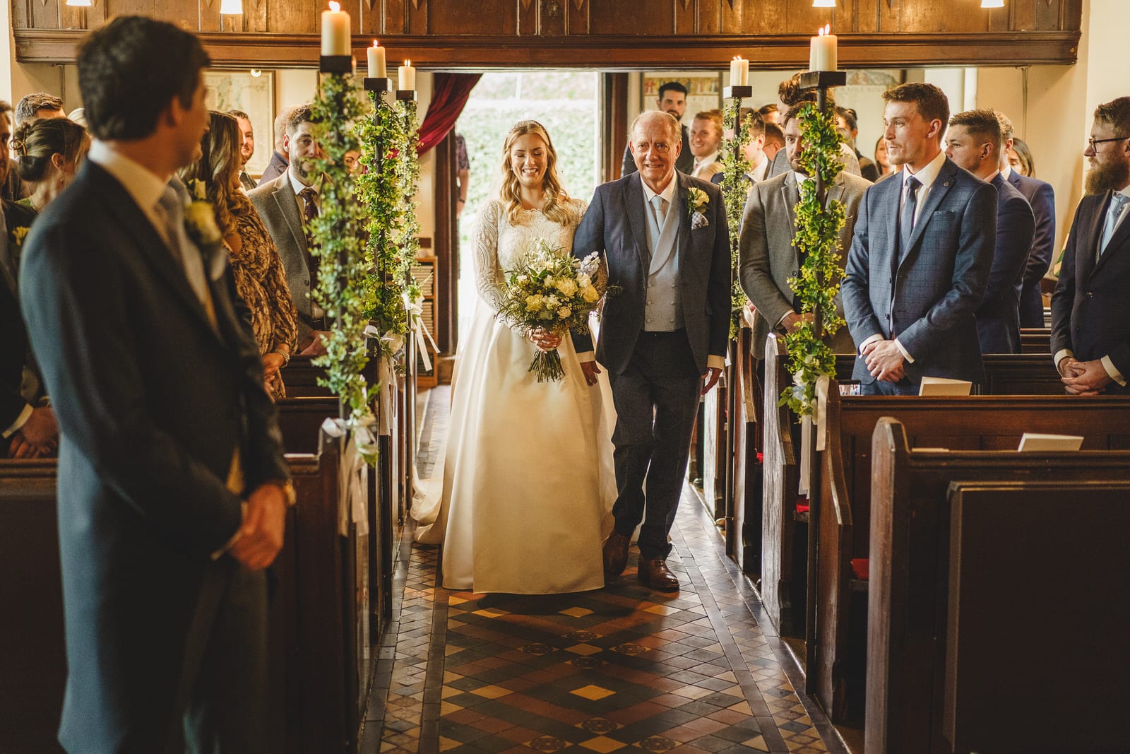 a bride in a white gown walks down the aisle with an older man in a church, surrounded by guests on both sides. it's a beautiful wedding scene in shropshire, capturing the heartfelt moment perfectly.