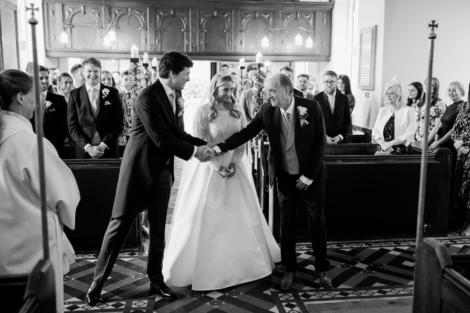 a bride stands between the groom, hamish, and an older man who are shaking hands inside a church filled with seated guests, capturing the moment of their charming wedding in Shropshire.