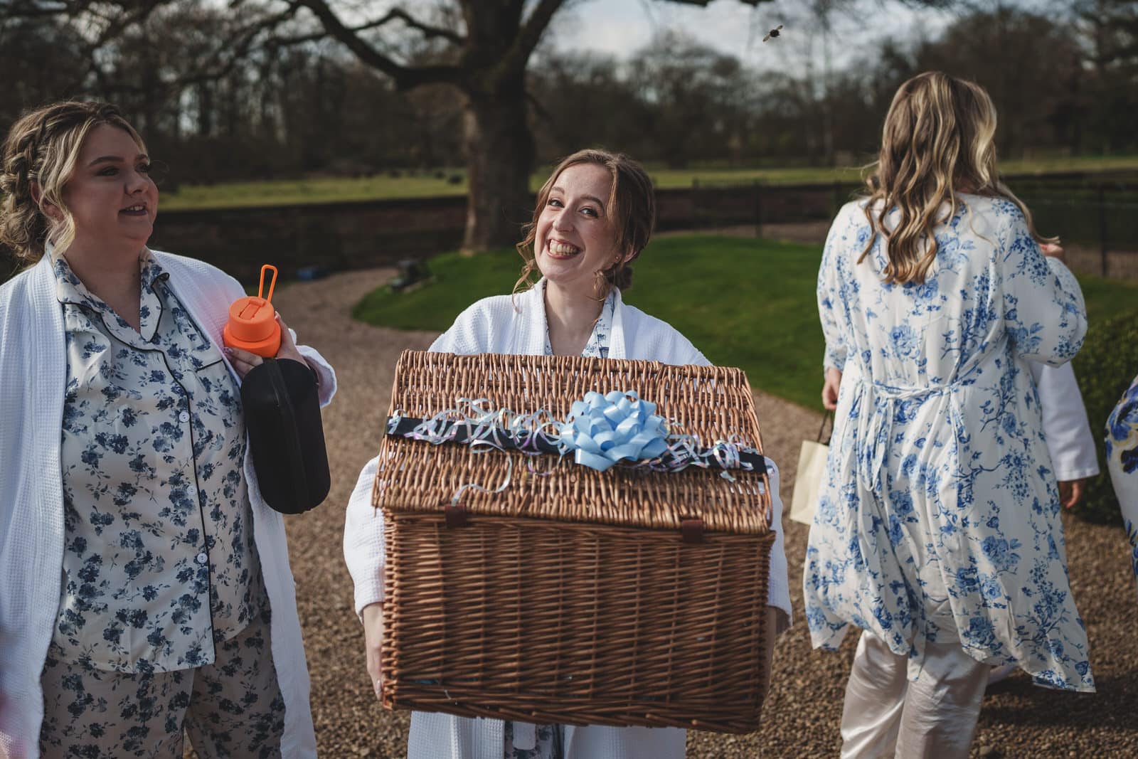 A person in a robe smiles while holding a large wicker basket with a blue ribbon at Iscoyd Park. Another robed individual holds a drink bottle, while a third person is seen from the back. The outdoor setting, surrounded by the picturesque trees of Shropshire, adds to the serene atmosphere.