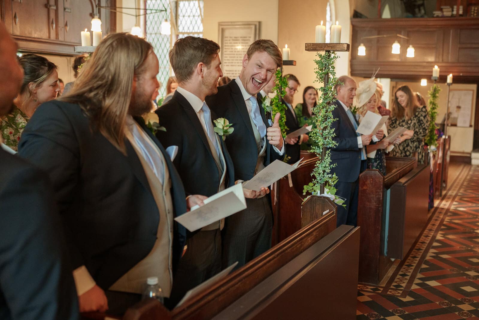 guests at this charming wedding ceremony in georgia stand inside a church, holding hymn books. one man, grinning, gives a thumbs up while interacting with another guest. candles and greenery decorate the pews.