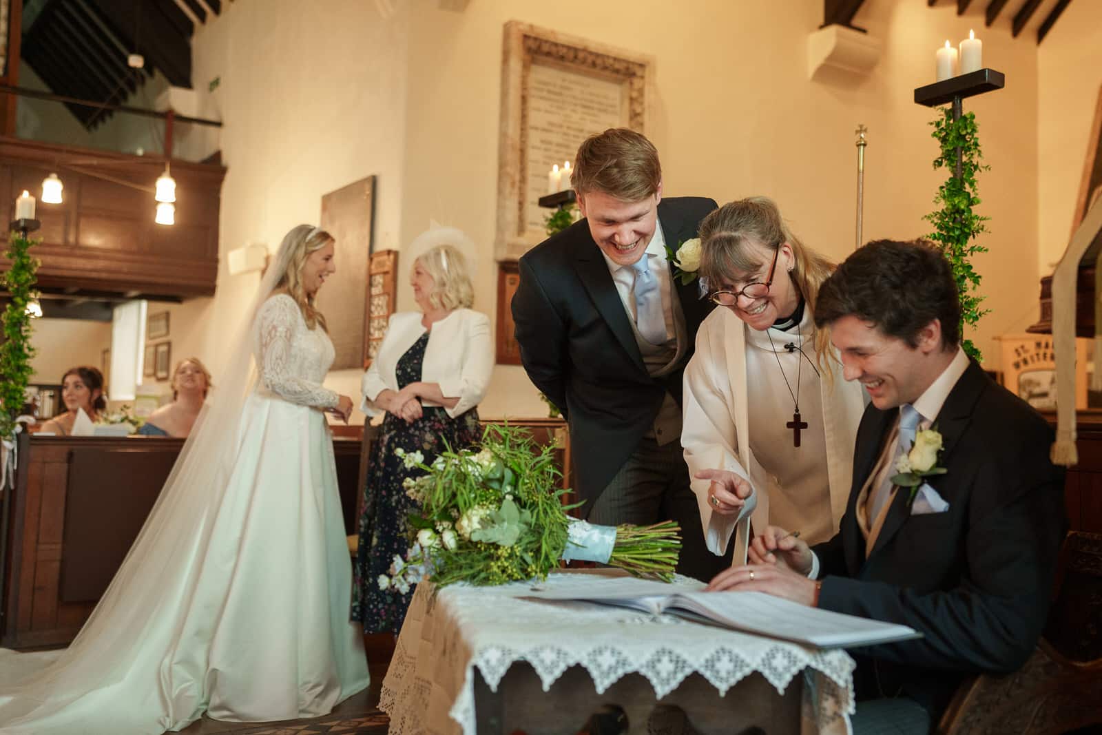a couple signs a wedding document inside a church with a priest and hamish, the groomsman, looking on. two women, one in a wedding dress, chat in the background near georgia. a floral arrangement adorns the table.