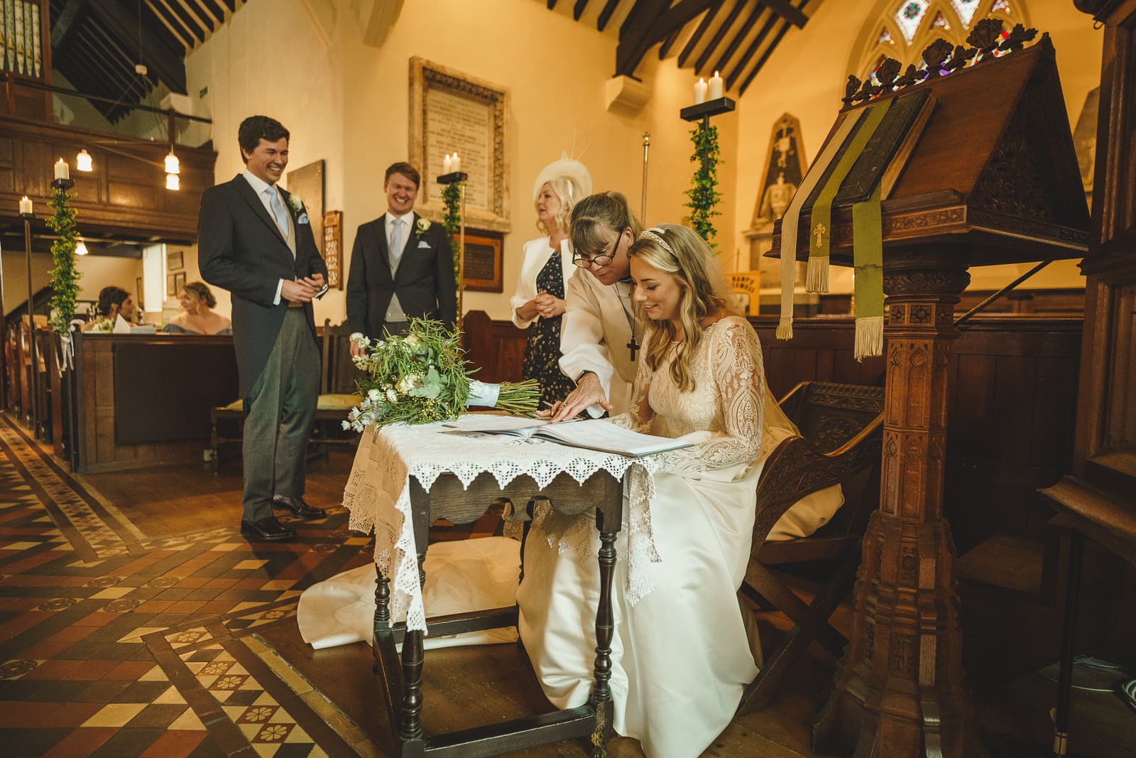 hamish and georgia sign marriage papers at a church altar, with three smiling guests standing nearby, all dressed in formal attire. the scene is warmly lit with candles and decorated with flowers, capturing the essence of their charming wedding.