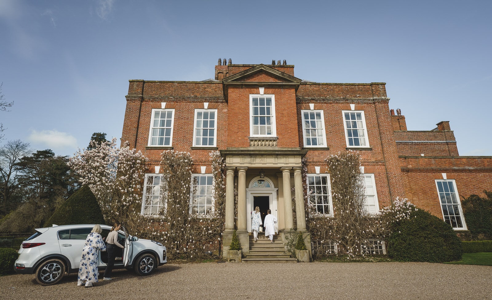 Three people stand near the entrance of a large brick mansion at Iscoyd Park in Shropshire, with a car parked in front. Two people are near the car, and one person is on the steps. The house has white-framed windows and charming climbing plants.