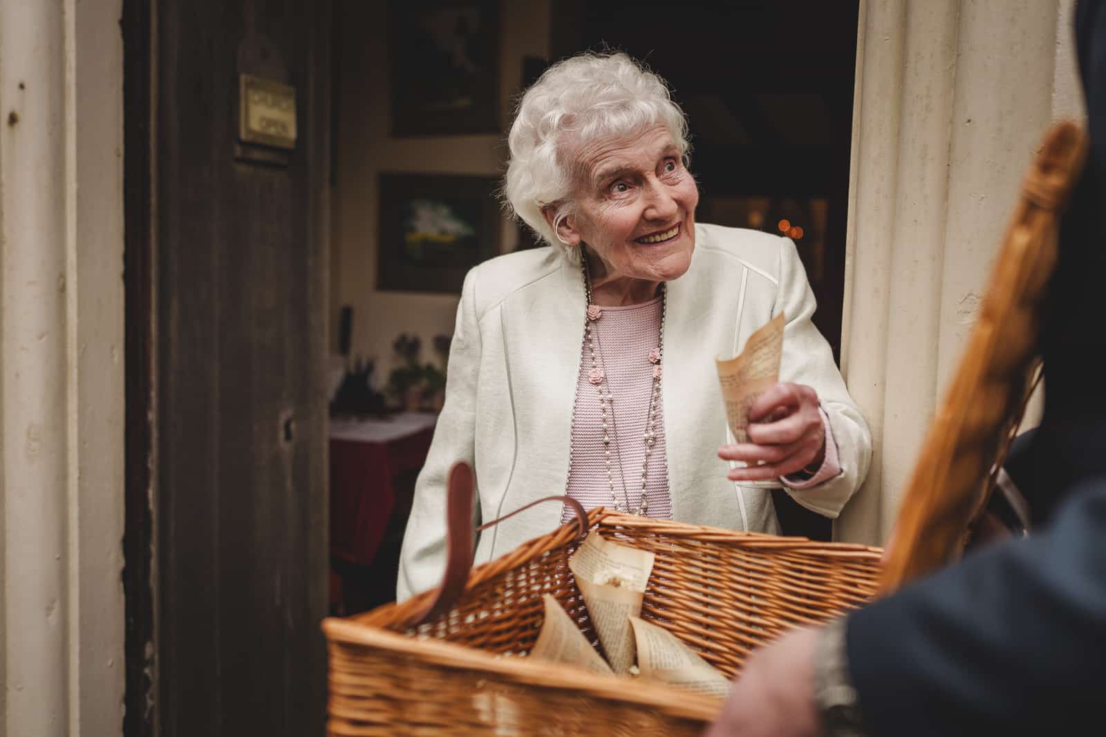 elderly woman with white hair and a white jacket smiles while holding a rolled up paper near a wicker basket. she stands in a doorway, perhaps reminiscing about georgia and hamish's charming wedding, engaging with someone out of the frame.