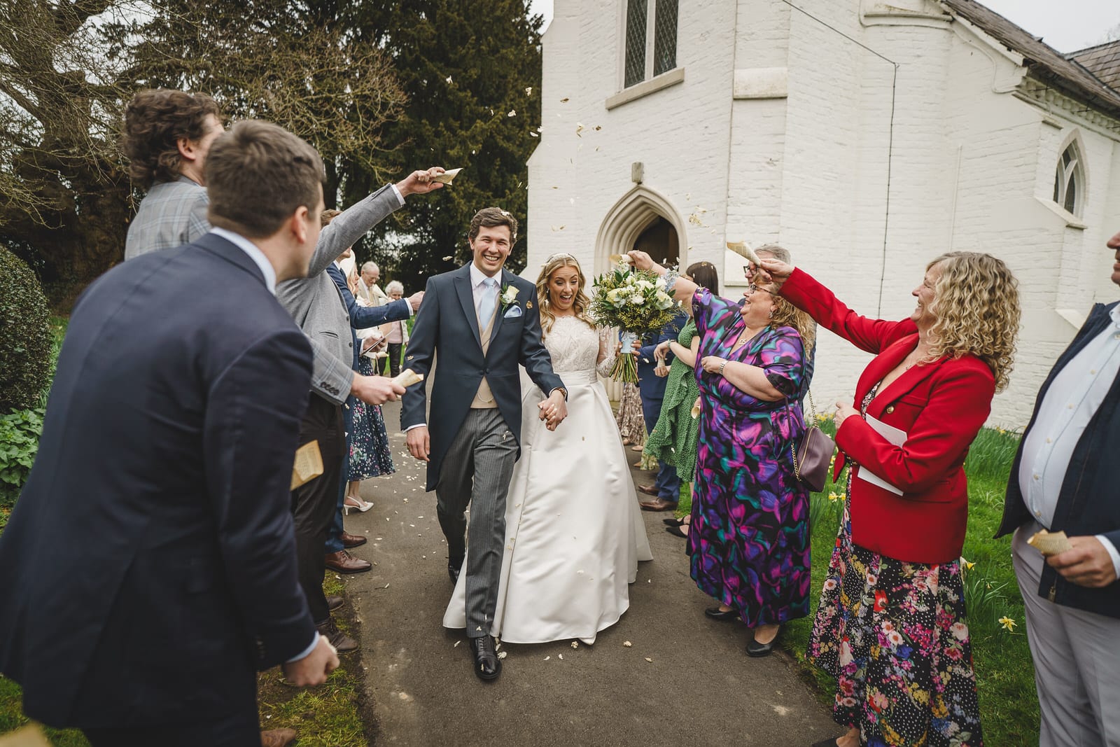 hamish and georgia, a charming newlywed couple, walk out of the church while guests throw flower petals at them. the bride is wearing a white gown, and the groom is in a dark suit with a bow tie.