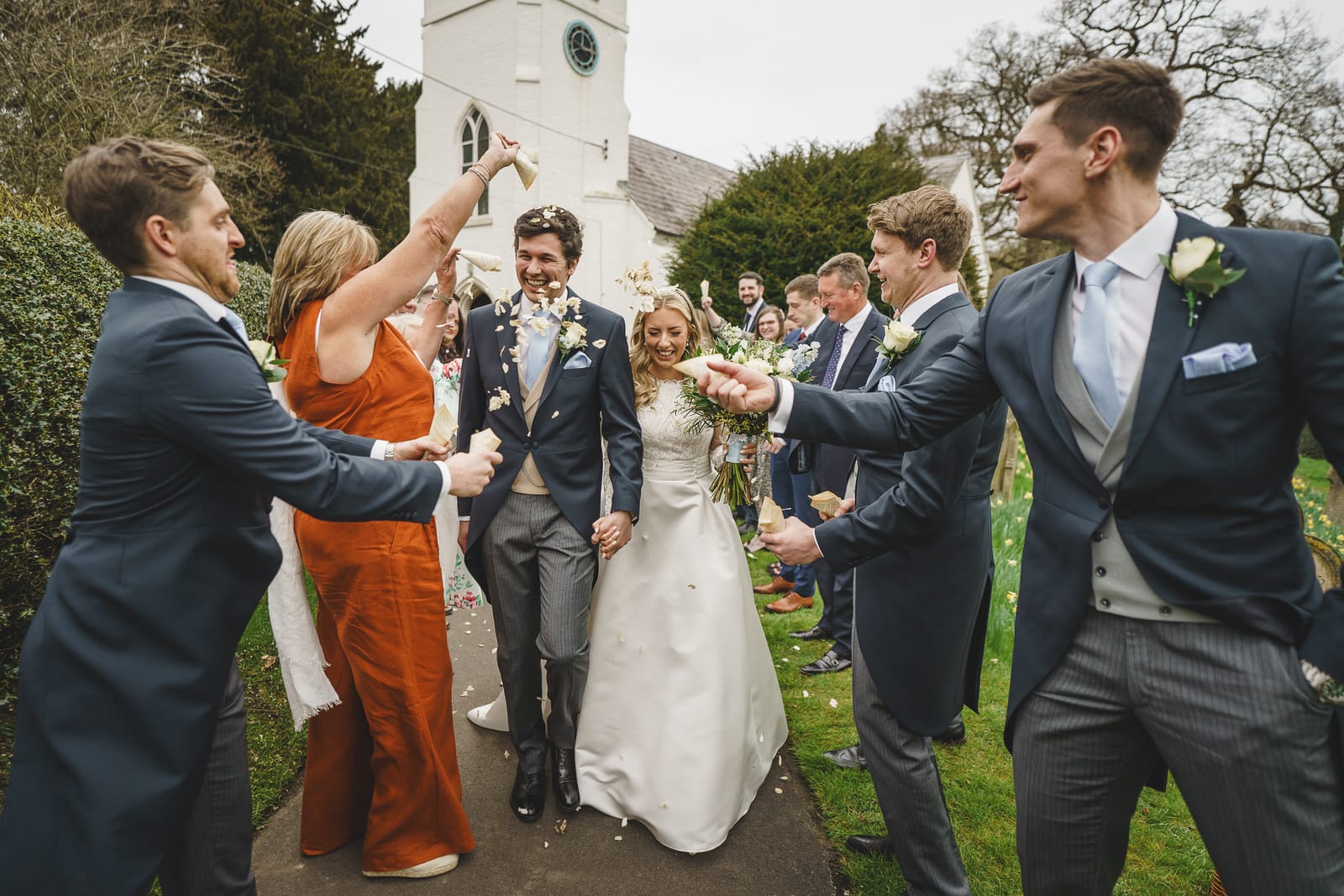 a bride and groom smile while exiting a charming wedding as friends and family, dressed in formal attire, cheer and throw confetti in celebration.