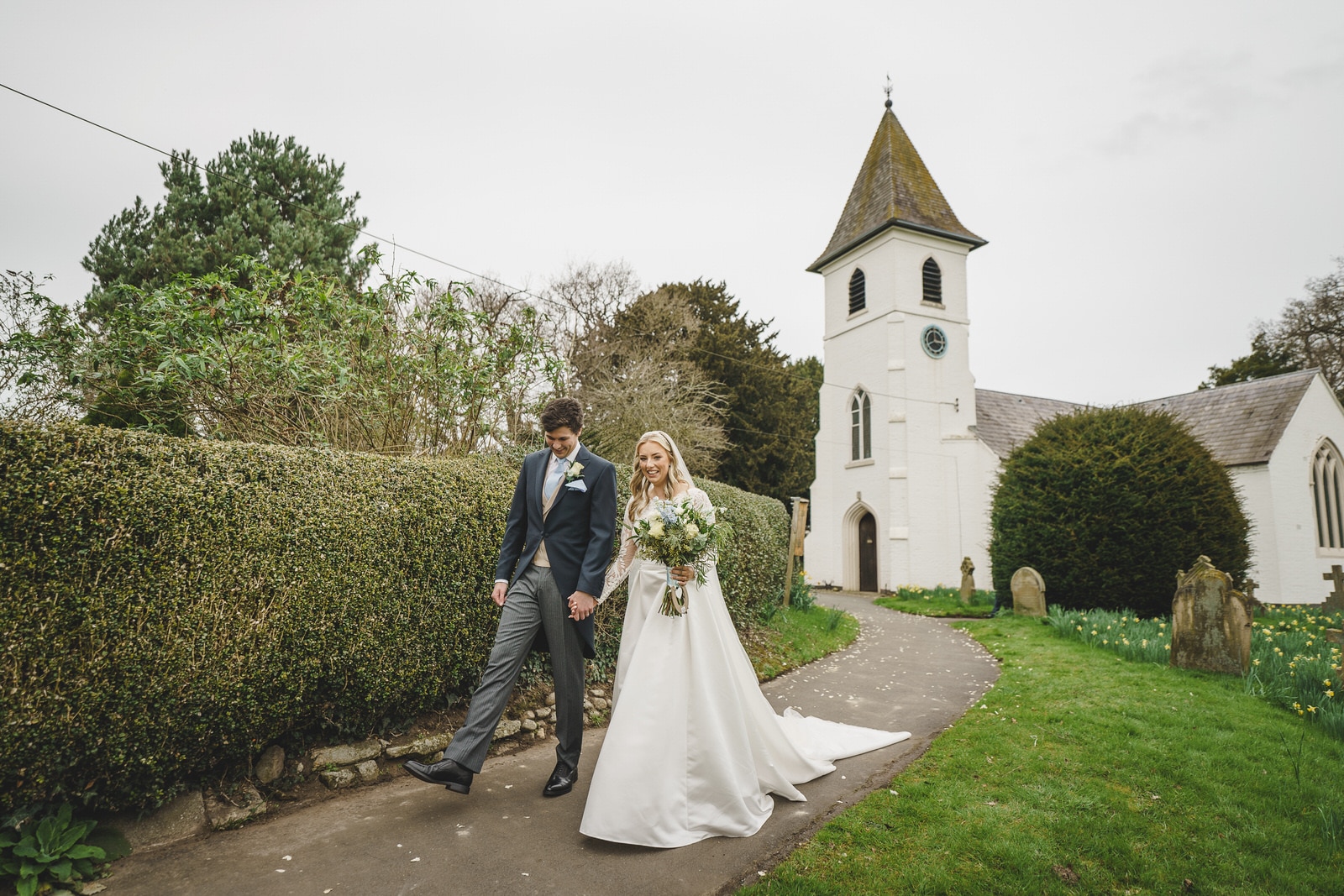 a newlywed couple, hamish and georgia, hold hands while walking outside a small, white church with a steeple. the bride is in a white gown, and the groom is in a suit. greenery and a path surround the charming wedding setting.