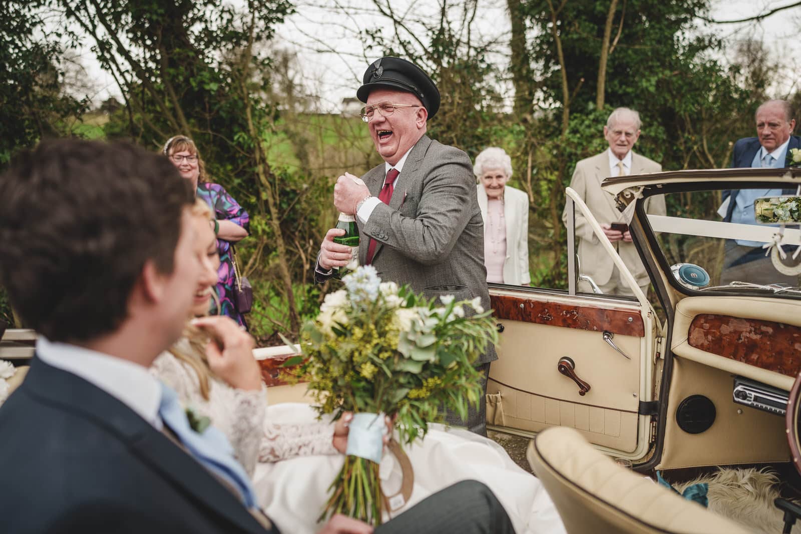 a man in a gray suit and hat laughs while holding a bottle of champagne next to a vintage car, with the charming wedding couple, hamish and georgia, sitting inside and guests smiling in the background.