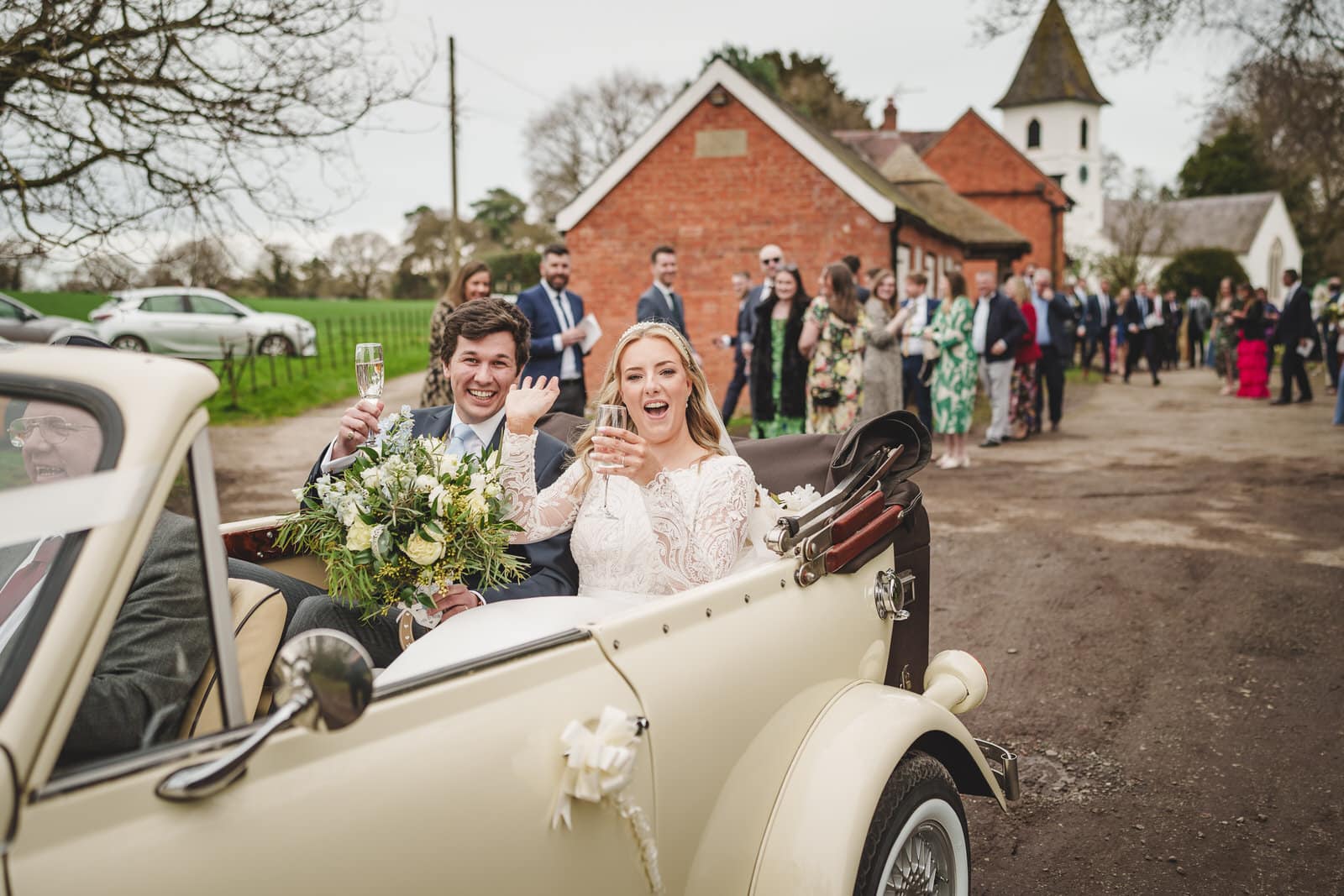 at the wedding of hamish and georgia, the bride and groom sit in a vintage convertible car holding glasses of champagne, with a group of well dressed people standing in the background near a brick building.