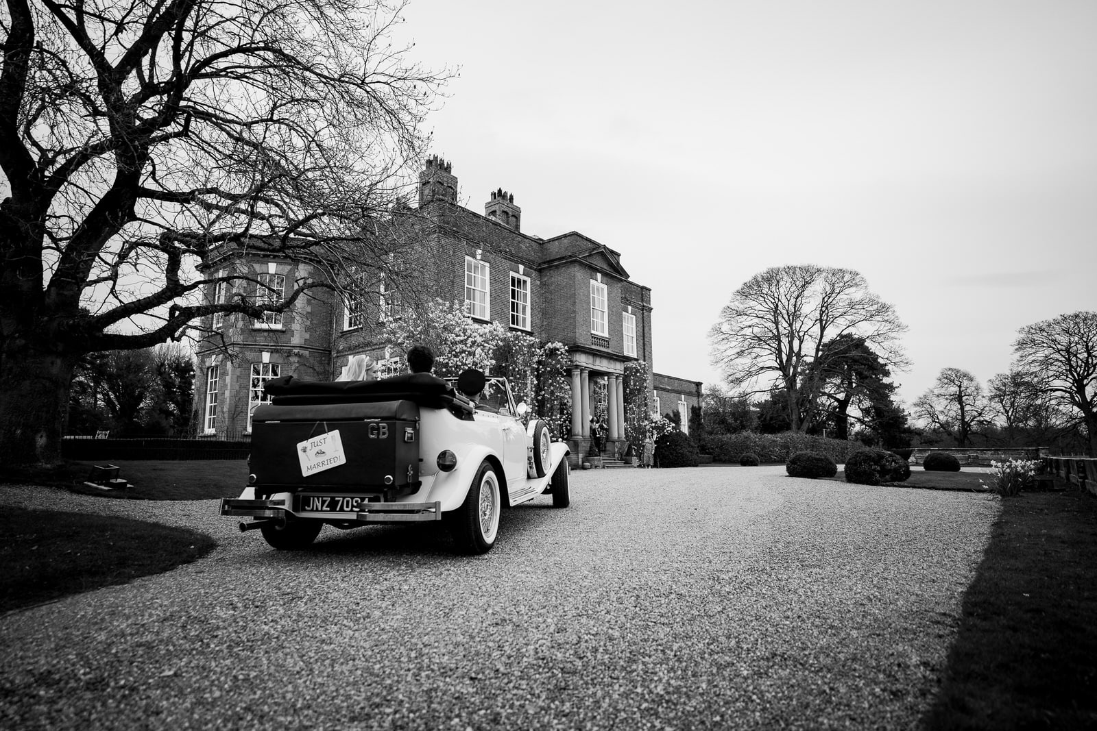 a vintage car, carrying hamish and georgia to their wedding, approaches a large, historic mansion with trees in the background, all captured in black and white.