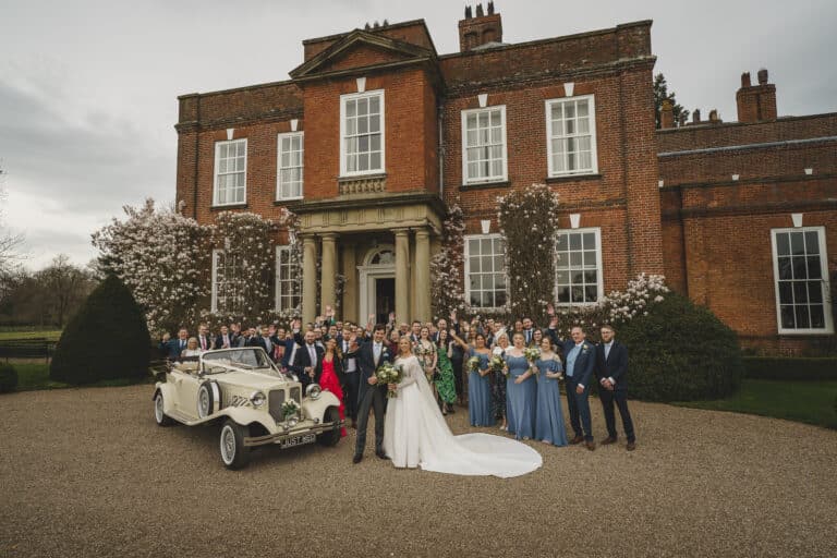 A large wedding party, including the bride and groom, pose in front of an elegant brick mansion at Iscoyd Park in Shropshire, with a vintage car parked nearby on a gravel driveway.