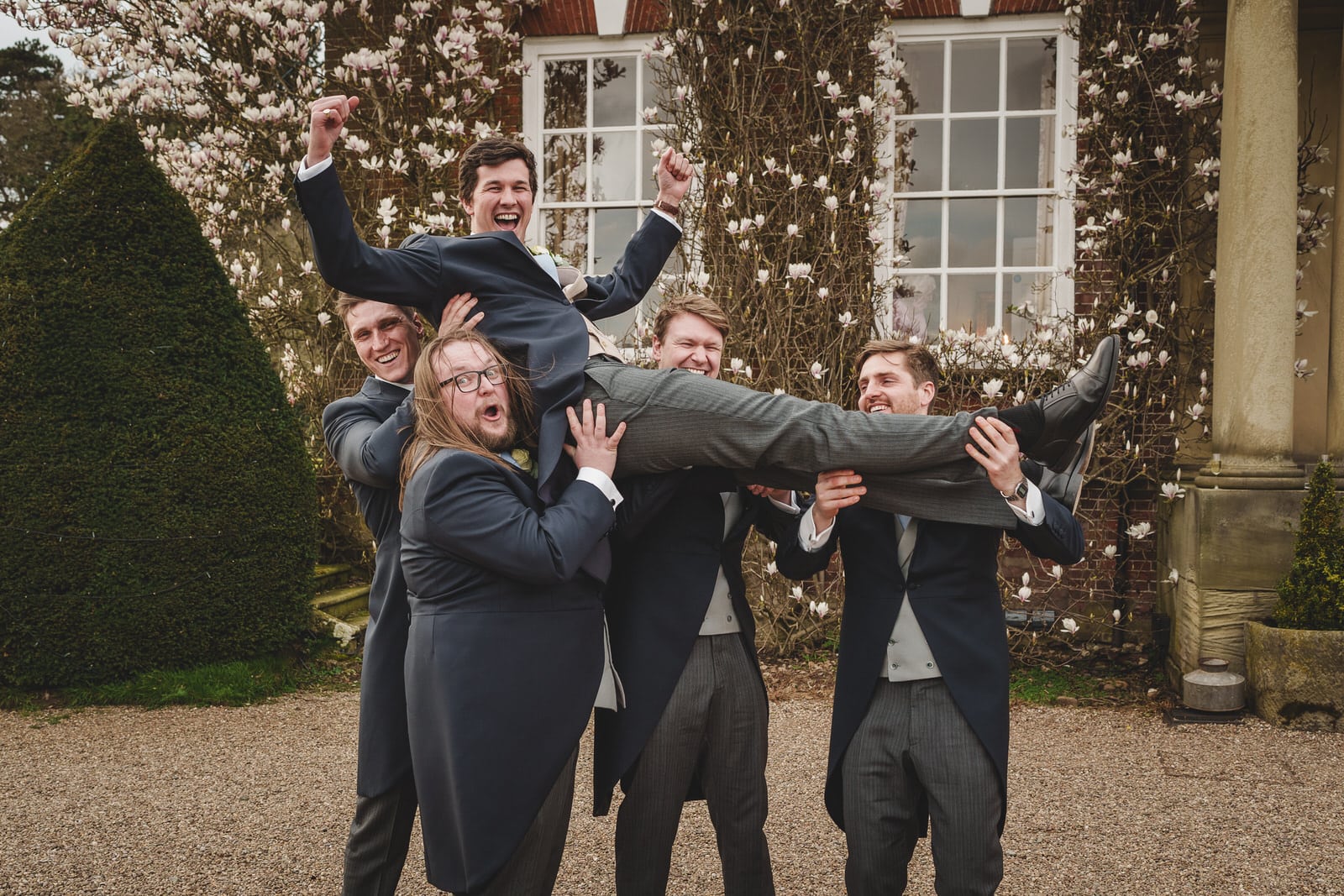 Five men in formal attire are outside a building with a flowering tree at Iscoyd Park. Four men are lifting the fifth man in the air as he raises his fists and smiles.
