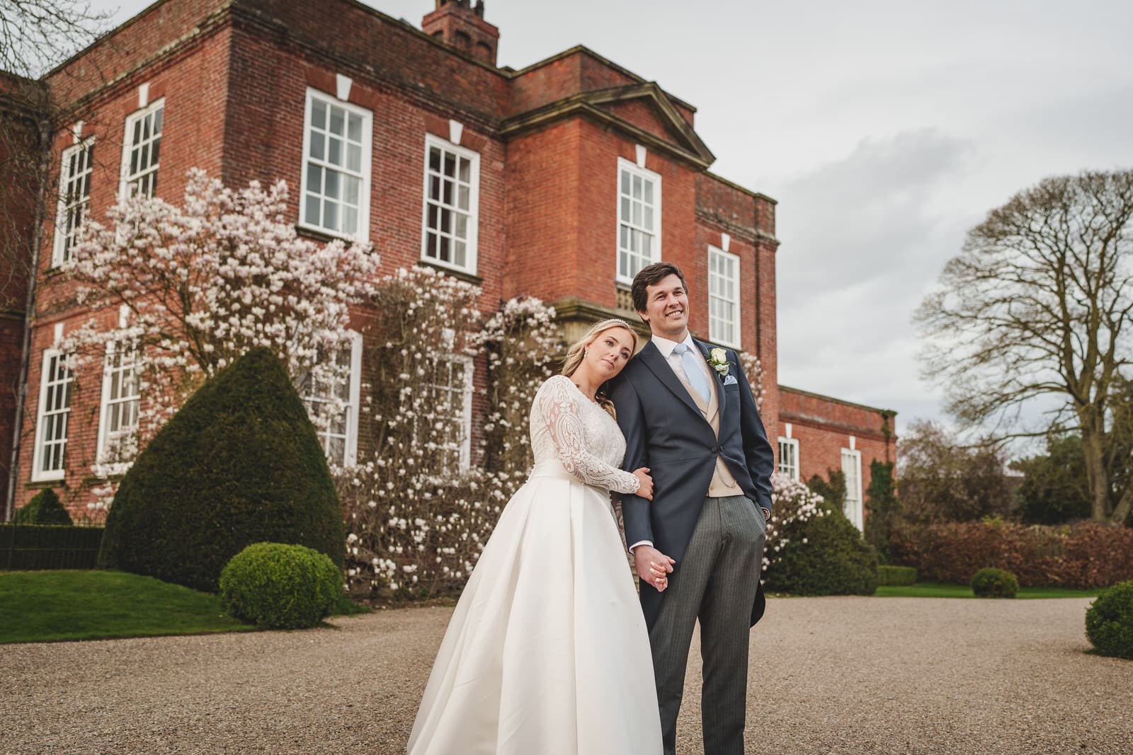 A bride and groom stand outside a large brick building with white-framed windows at Iscoyd Park in Shropshire. The bride is wearing a white dress, and the groom is in a suit. They are holding hands and looking at the camera.