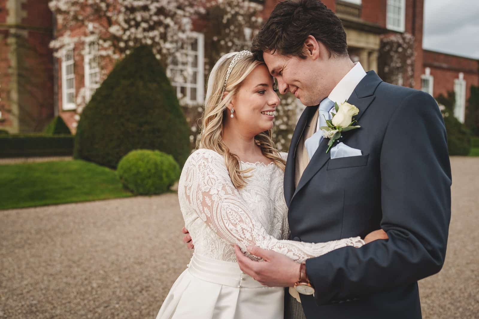 A bride and groom embrace and smile at each other outdoors in front of the large building at Iscoyd Park, Shropshire, with manicured bushes and a blossom tree in the background.