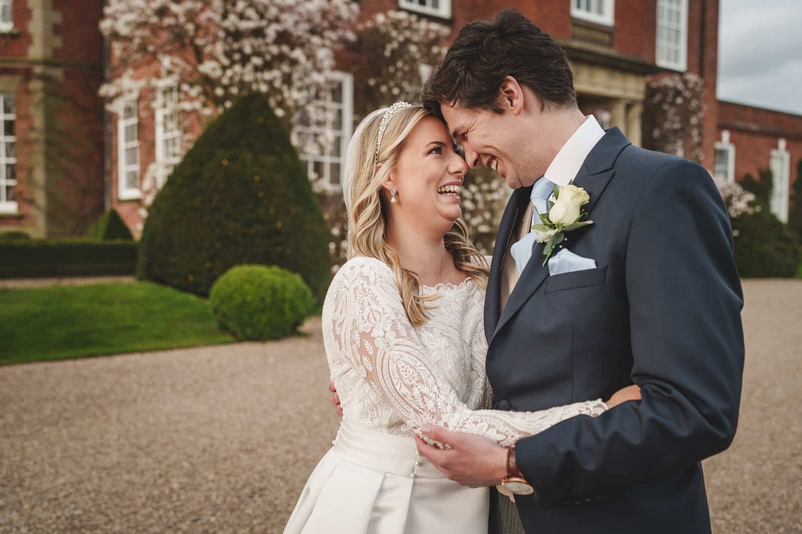 Hamish and Georgia laugh and embrace outside a large brick building, its manicured gardens providing the perfect backdrop for their charming wedding.