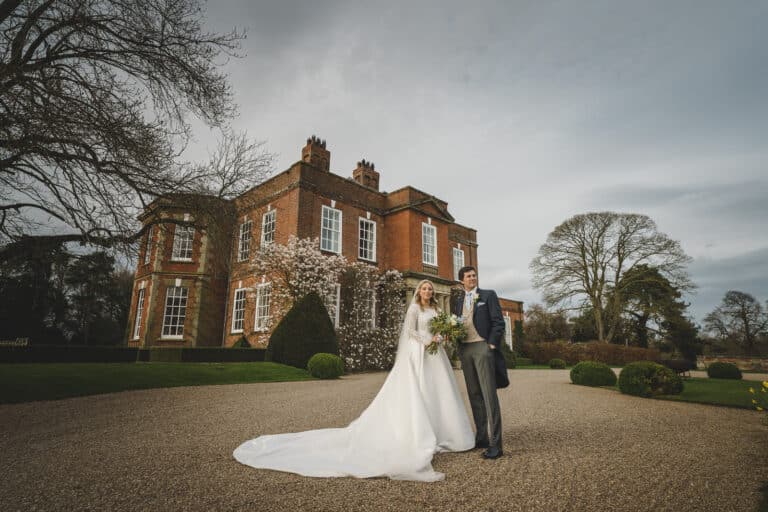 A couple dressed in wedding attire stands together outside a large brick building, surrounded by a gravel pathway and manicured bushes. The bride, Georgia, holds a bouquet, and both she and Hamish are smiling.