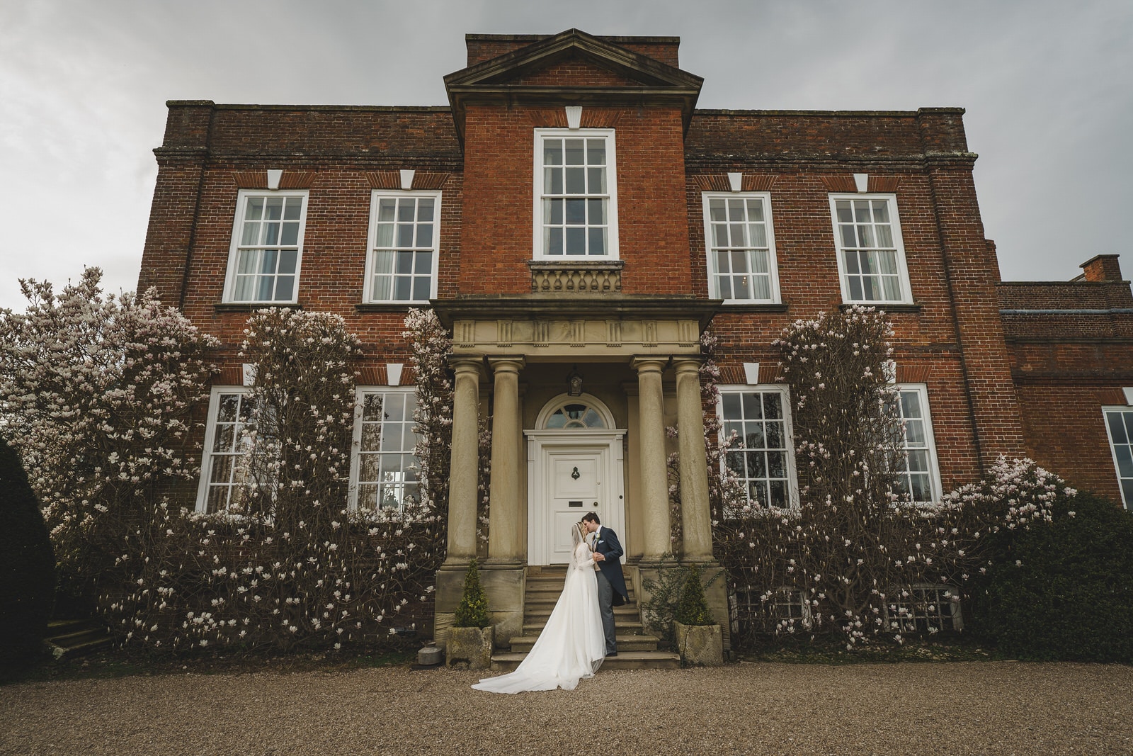 Hamish and Georgia stand on the steps in front of a large, red-brick building adorned with white blossoming bushes, capturing their charming wedding day.