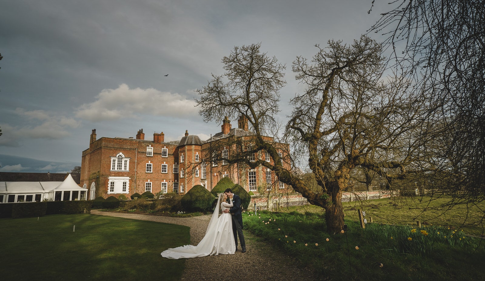 Hamish and Georgia stand in front of a grand brick mansion on a cloudy day, with trees and a manicured lawn surrounding them, creating the perfect backdrop for their charming wedding.