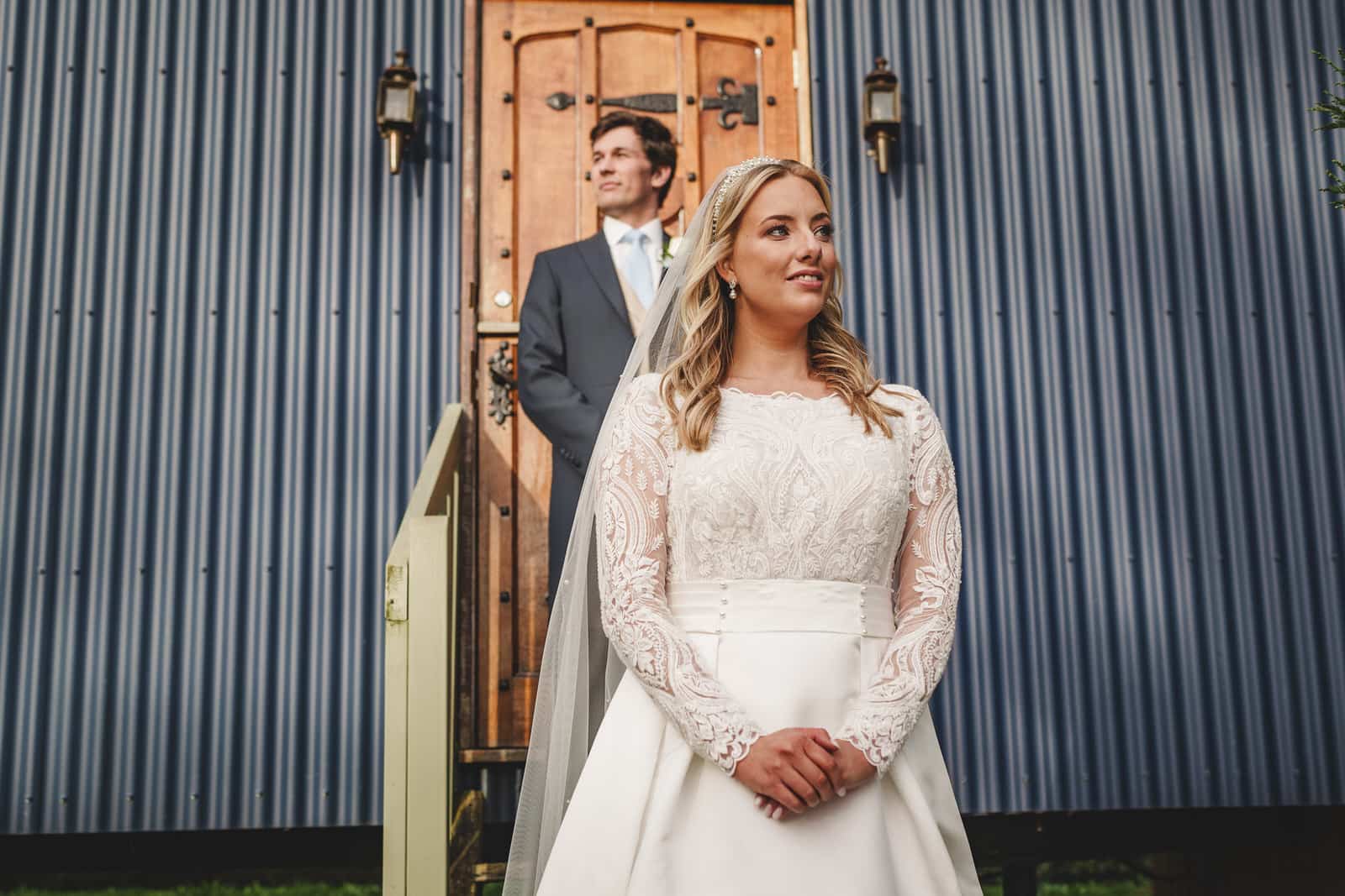 A bride in a white dress stands in front of a groom in a suit, celebrating their charming wedding against the backdrop of a wooden door with metal siding in Georgia.