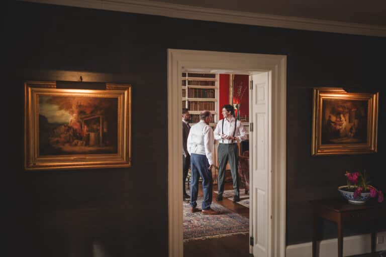 Three men are talking inside a room with bookshelves and artwork; seen through a doorway, there are paintings on both walls flanking the entrance, hinting at the timeless elegance of a Shropshire manor like Iscoyd Park.