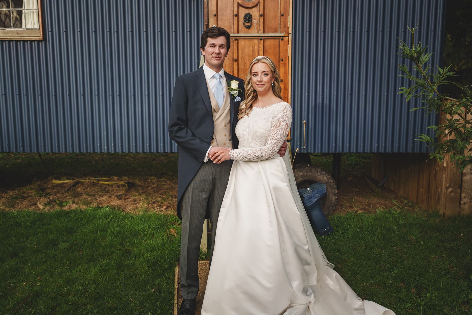 A bride and groom stand together outdoors in front of a blue wooden structure at Iscoyd Park. The bride is wearing a white wedding dress with lace sleeves, and the groom is in a dark suit with a cream-colored waistcoat, epitomizing an elegant Shropshire wedding.