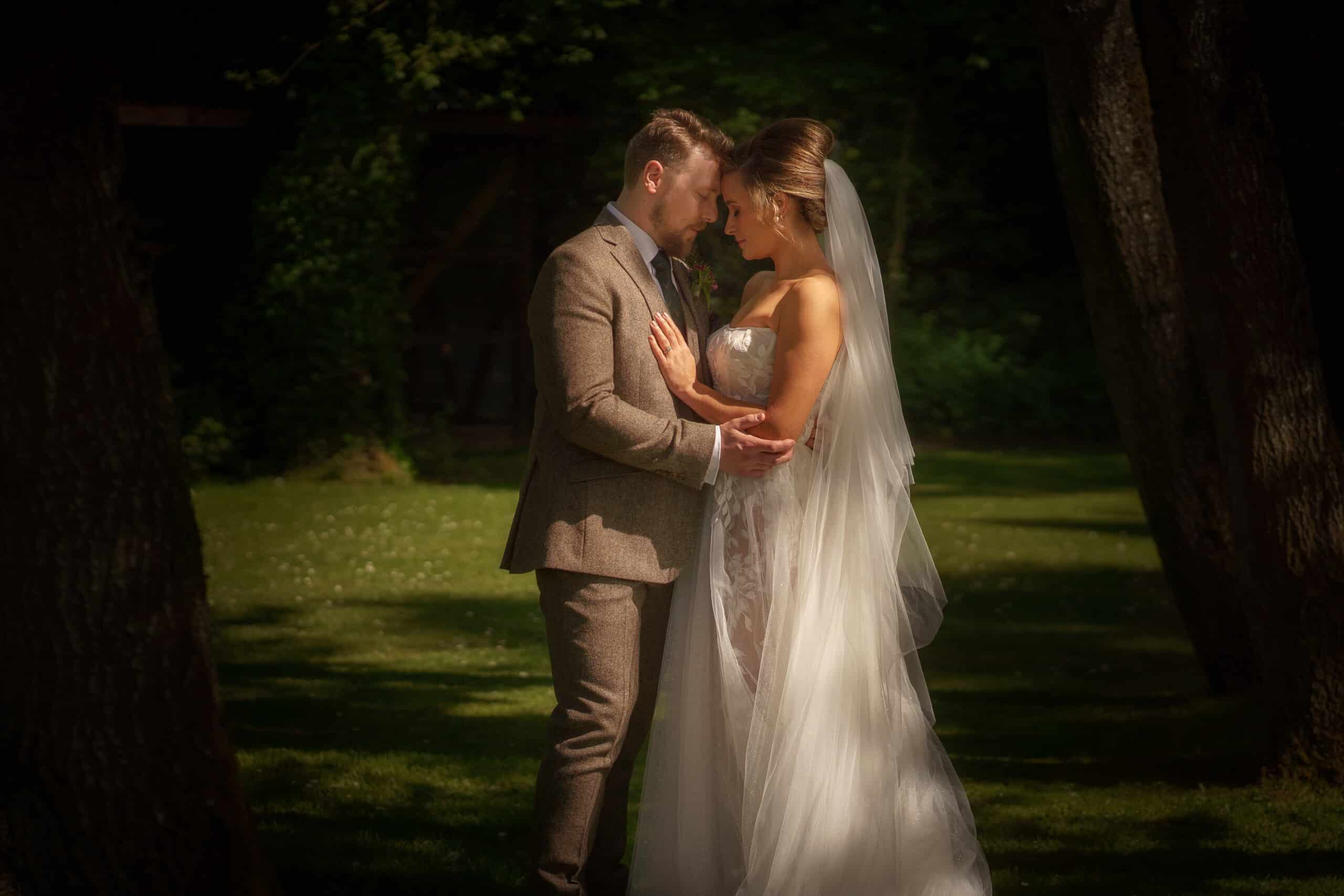 a bride and groom stand closely together outdoors, with the bride in a white gown and veil and the groom in a brown suit, surrounded by trees and greenery, captured perfectly by a wedding photographer in shropshire.