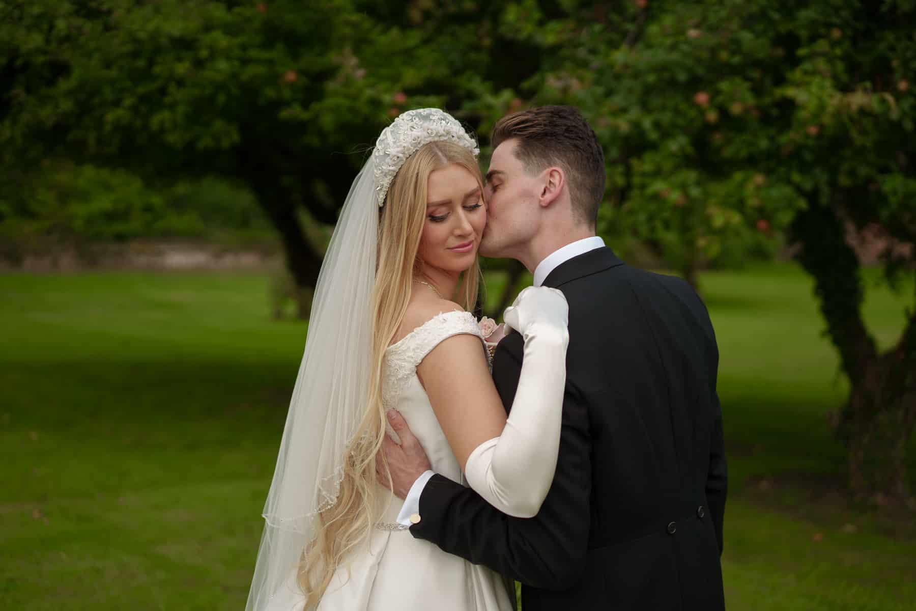 a bride and groom embrace outdoors, with the groom leaning in to kiss the bride's cheek. the bride wears a white dress and veil, while the groom is in a black suit. greenery is in the background, perfectly captured by pbartworks photography, renowned wedding photographers in shropshire.