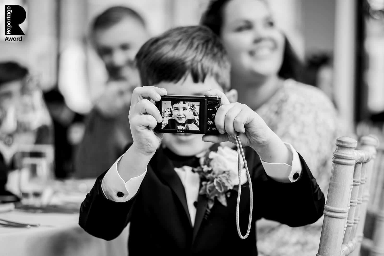 a child dressed in a suit holds a camera, taking a photo. the camera screen displays the child's face. two adults are blurred in the background, sitting at a table — capturing timeless moments with pbartworks photography.