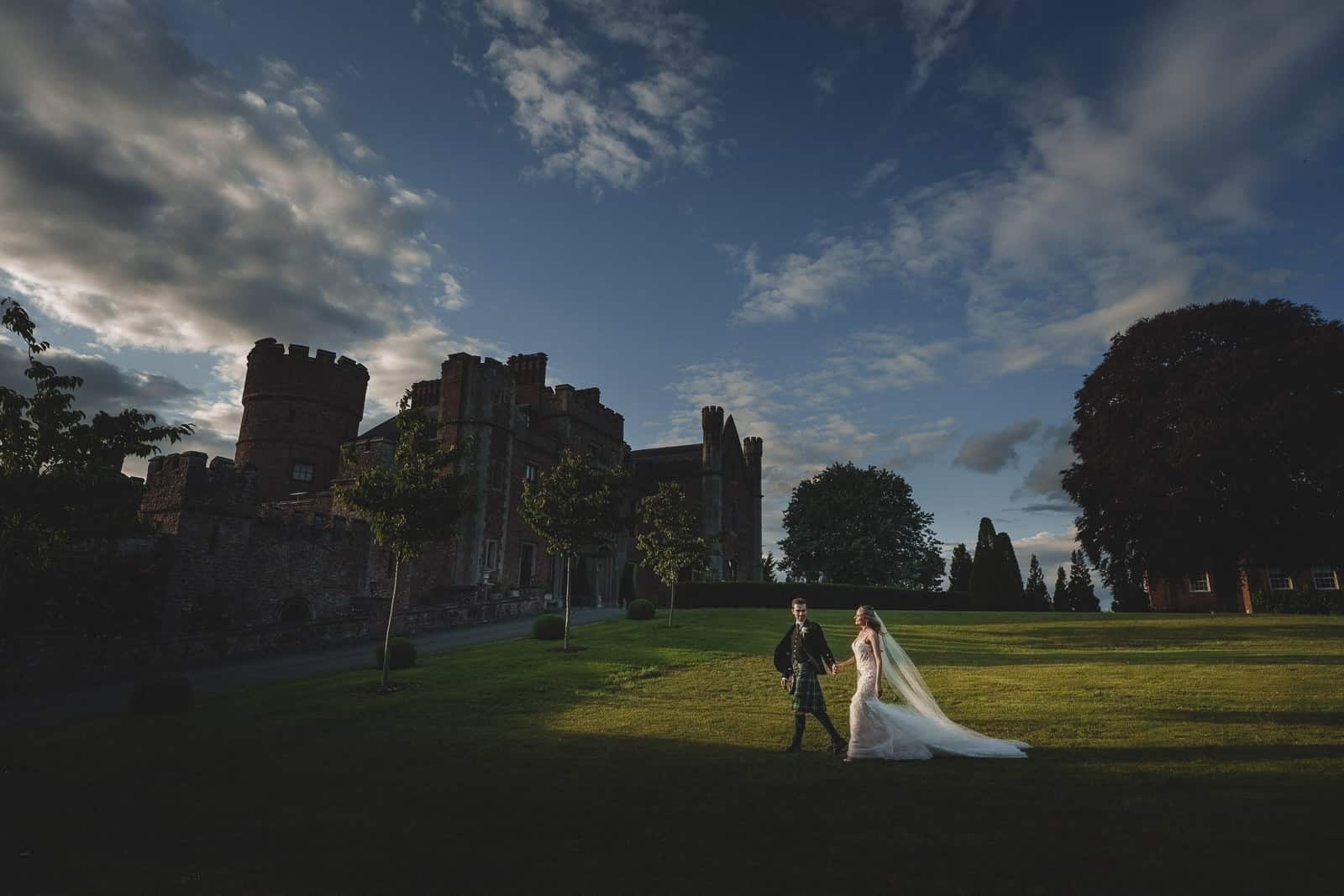 a bride and groom walk across a sunlit lawn in front of a large castle under a partly cloudy sky, beautifully captured by pbartworks photography, one of the leading wedding photographers in shropshire.